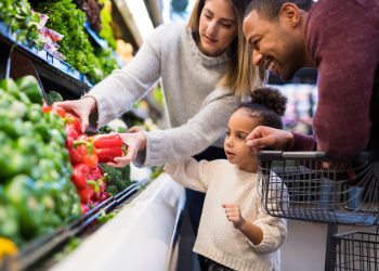 A pre-school age girl helps her parents pick out veggies in the produce section at the grocery store. She is reaching for a red pepper.