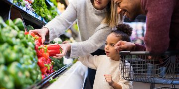 A pre-school age girl helps her parents pick out veggies in the produce section at the grocery store. She is reaching for a red pepper.